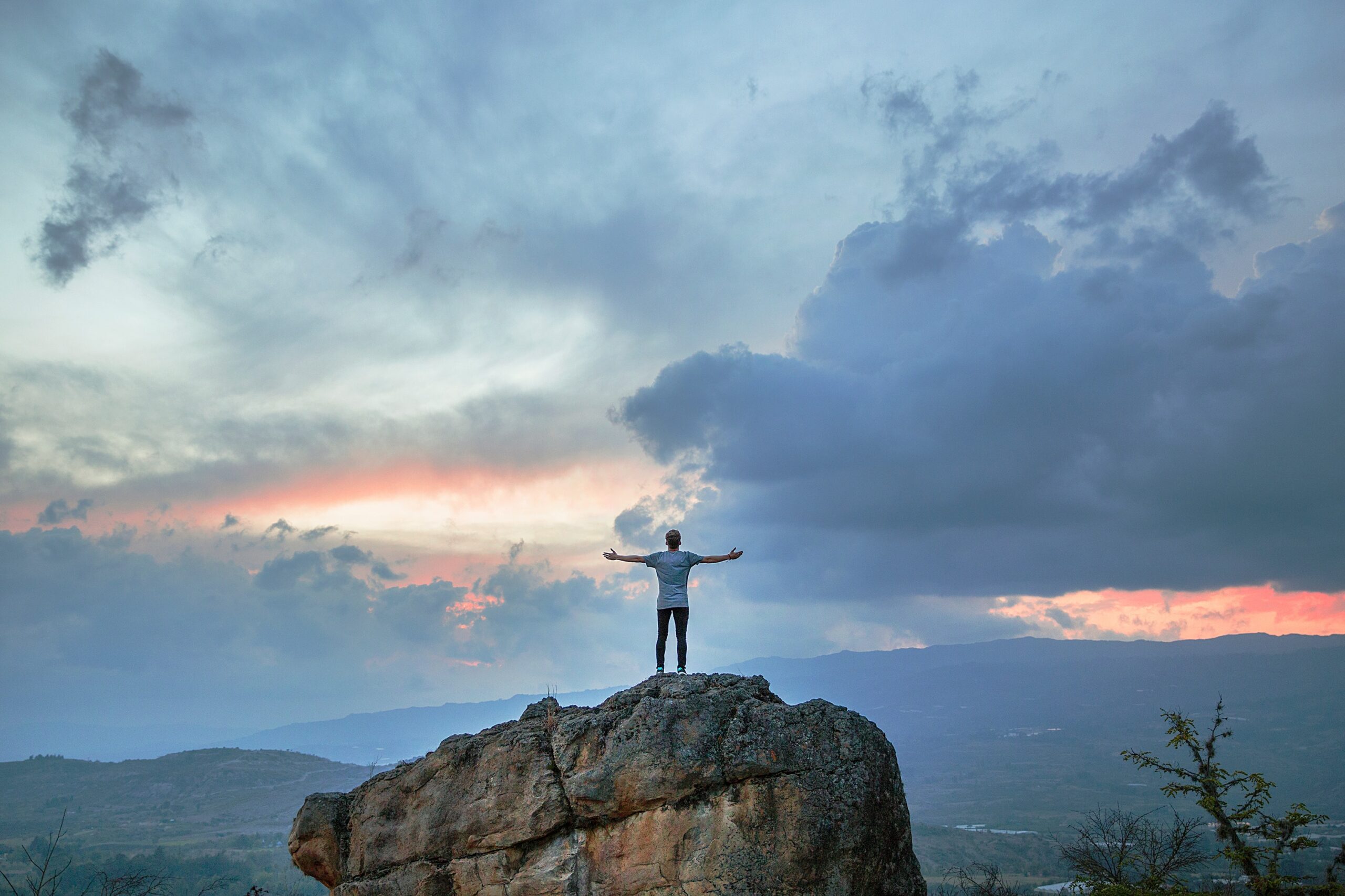Man standing on a large rock with his arms open, facing a sunrise.